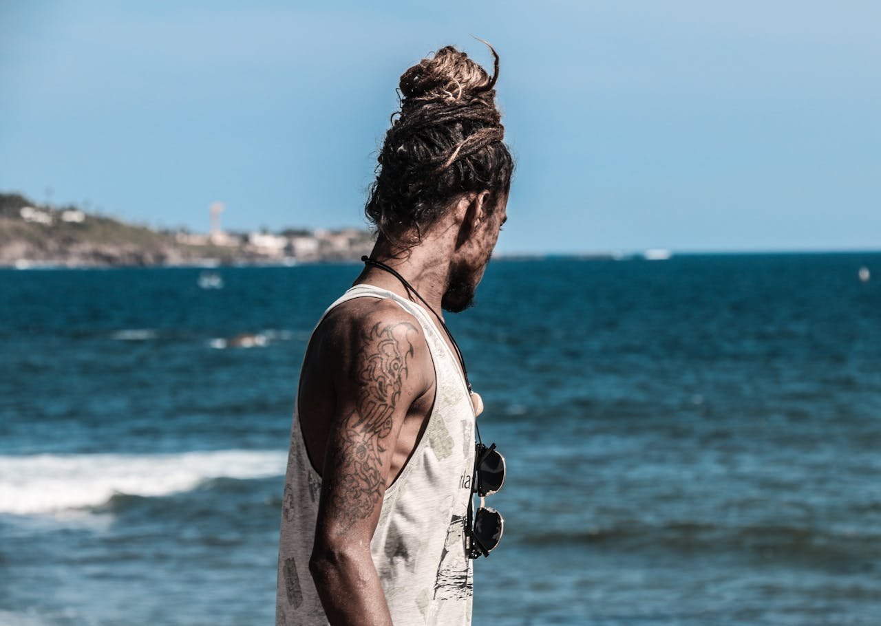 A tattooed man with dreadlocks enjoying a sunny day at the beach overlooking the ocean.