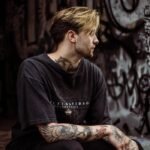 A tattooed man in a black T-shirt sits in a graffiti-covered urban environment, showcasing street style.