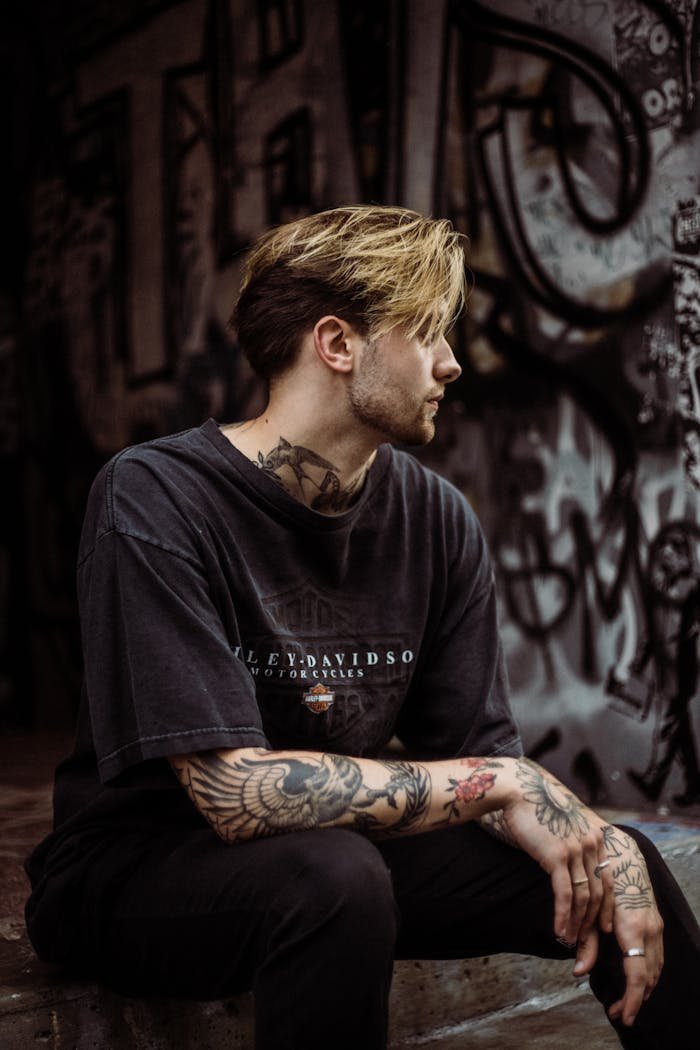 A tattooed man in a black T-shirt sits in a graffiti-covered urban environment, showcasing street style.