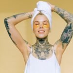 Tattooed woman enjoying spa day indoors with gold eye patches and towel on head.