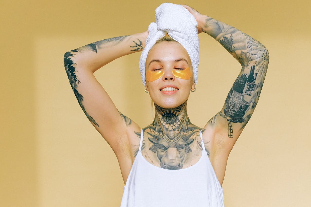Tattooed woman enjoying spa day indoors with gold eye patches and towel on head.