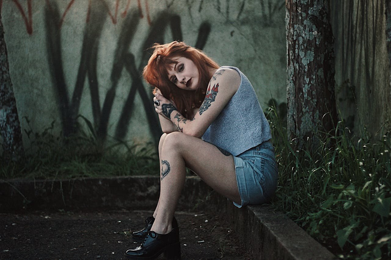 Stylish redhead woman with tattoos posing outdoors with graffiti backdrop.